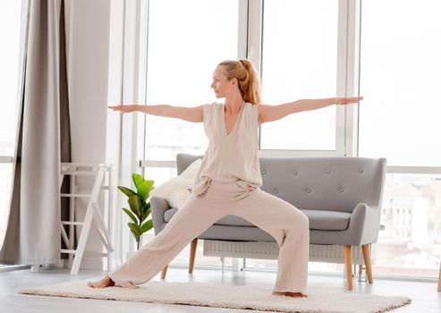 Blond woman girl practicing yoga at home. Stretching workout in room with sunlight