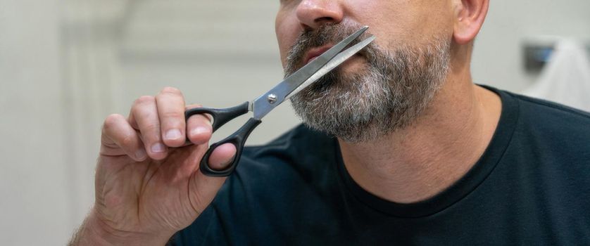 Portrait of Middle-aged handsome man cutting his beard with scissors. Selfcare during quarantine isolation