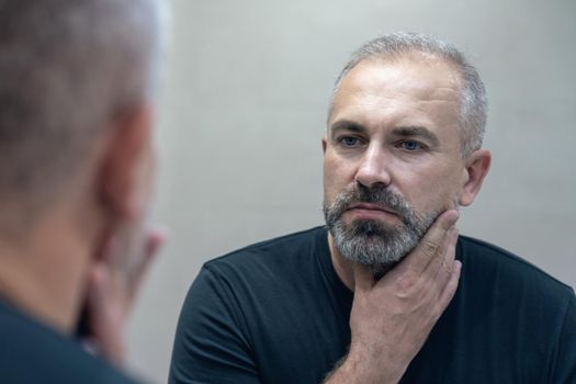 Middle-aged handsome man looking in mirror in bathroom touching his beard thinking of cutting it off