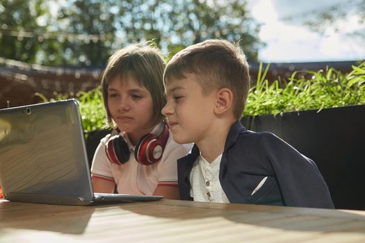 bored little caucasian schoolchildren sit outdoors, watch at screen of laptop
