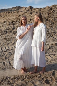 two attractive young twin sisters posing in sand quarry in elegant white clothes