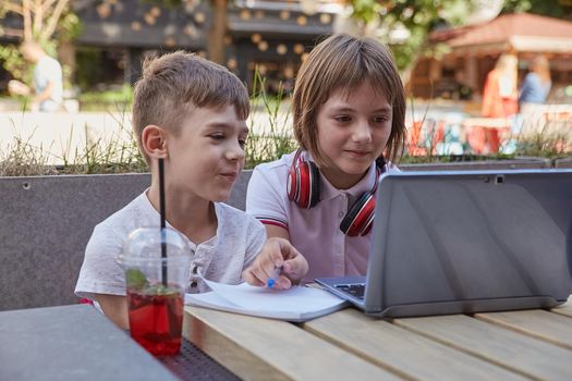 little caucasian schoolchildren sit outdoor, watch at screen of laptop, have fun