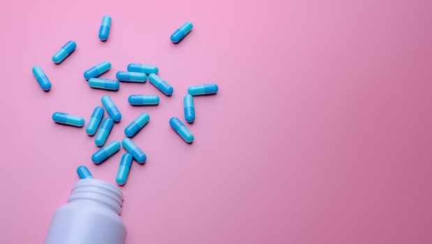 Blue capsule pills spread out of white plastic drug bottle on pink background. Pharmacy banner. Online pharmacy. Painkiller medicine and antibiotic drug resistance concept. Pharmaceutical industry.