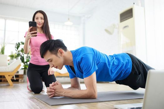 Smiling young woman with smartphone and man exercising and doing plank at home.