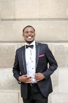 Afro american successful smiling man wearing dark suit and standing near wall outdoors.