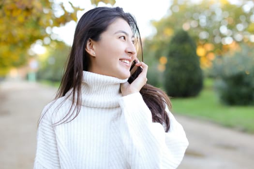 Asian happy girl wearing white sweater talking by smartphone and walking in park.