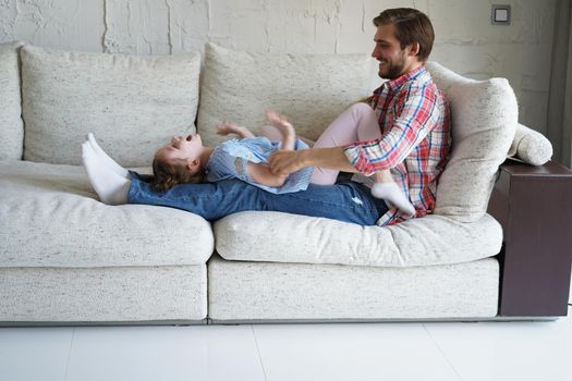 Cute little kid daughter laughing tickling playing with dad on sofa, happy father relaxing having fun with funny small child girl bonding enjoying leisure together in living room.