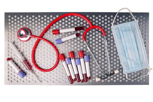 Flat lay of blood sample along with syringe, protective mask and stethoscope on metal surface