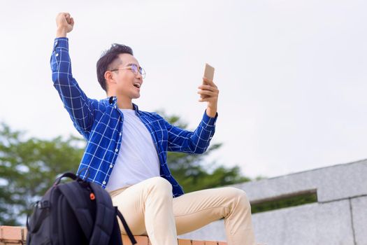excited young asian man sitting on stairs and using smart phone