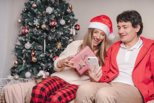 Portrait of lovely couple husband and wife with mobile phone in red Santa hat having a video call chat with puppy dog in sweater, enjoy Christmas time at home. Online greeting and taking selfie.