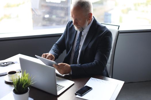 Mature business man in full suit using digital tablet while sitting in the office.