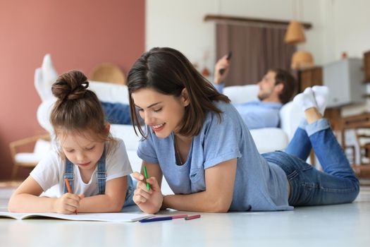 Happy mother smilling daughter lying on warm floor enjoying creative activity, drawing pencils coloring pictures in albums, father resting on couch, family spend free time together.