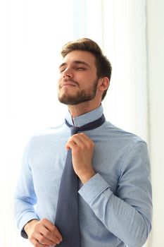 Morning dress up. Handsome young man in blue shirt adjusting his necktie and looking away.