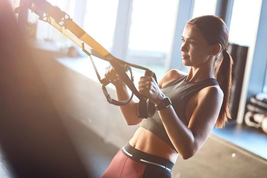 Side view of a young strong and fit woman exercising with fitness trx straps at gym