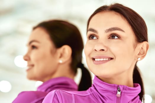 Close up portrait of a young beautiful caucasian woman, female fitness instructor in sportswear looking away and smiling while standing in studio or gym against mirror. Sport, wellness and healthy lifestyle