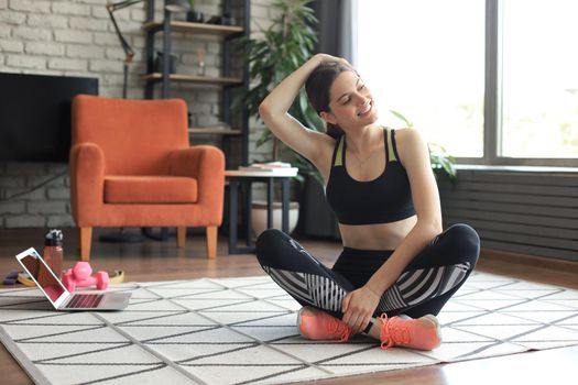 Fitness beautiful slim woman is sitting on the floor with dumbbells and bottle of water using laptop at home in the living room. Stay at home activities.