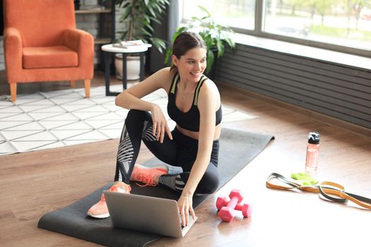 Beautiful slim sporty woman in sportswear is sitting on the floor with dumbbells and bottle of water and is using a laptop at home in the living room. Healthy lifestyle. Stay at home activities.
