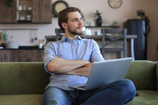 Concentrated young freelancer businessman sitting on sofa with laptop, working remotely online at home.
