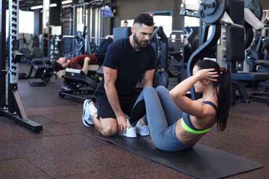 Trainer helping young woman to do abdominal exercises in gym.
