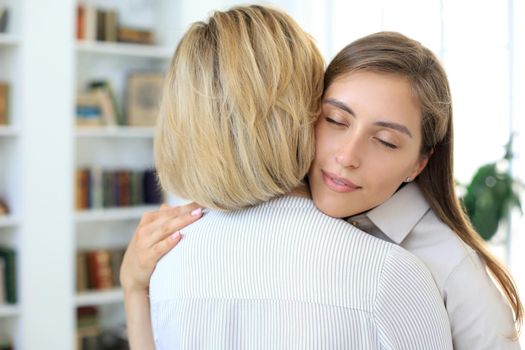 Cheerful young woman is embracing her middle aged mother in living room.