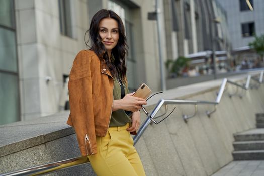 Side view of a young beautiful and fashionable woman holding her smartphone looking aside and smiling while standing on the city street, business lady walking outdoors