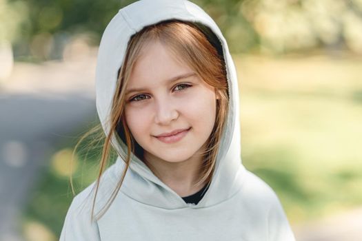Portrait of happy little girl in hoodie smiling at camera while walking in park on blurred nature background