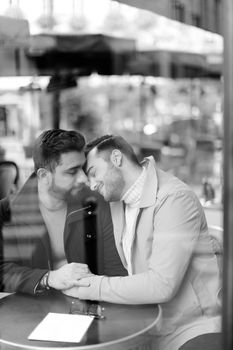 Black and white photo of caucasian gays sitting at cafe and hugging, reflection in glass.