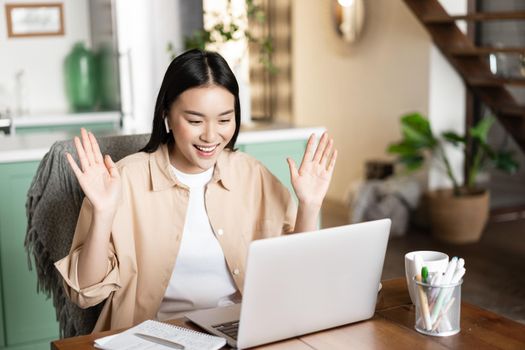 Asian girl on video conference, saying hello, waving hand at laptop camera, working remote from home. Young woman connects to webinar or online classes