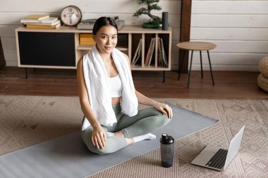 Asian woman doing morning yoga at home, sitting on mat in meditation pose, wearing activewear leggings and smiling peaceful