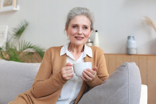 Happy mature woman resting on comfortable sofa drink coffee or tea, looking away, relaxing on cozy couch at home enjoy hot beverage