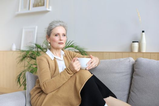 Happy mature woman resting on comfortable sofa drink coffee or tea, looking away, relaxing on cozy couch at home enjoy hot beverage