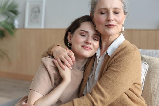 Portrait of old mother and mature daughter hugging at home. Happy senior mom and adult daughter embracing with love on sofa.