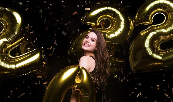 Happy gorgeous girl in stylish sexy party dress holding gold 2022 balloons, having fun at Christmas or New Year's Eve Party.