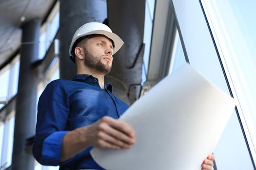 Confident young businessman in hardhat holding blueprint and inspecting it while standing indoors.