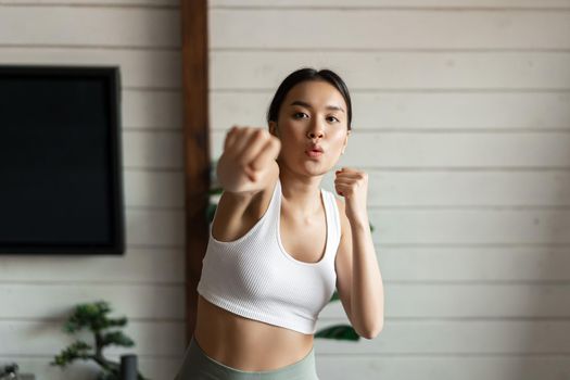 Fitness girl doing her workout at home. Asian athletic woman practice punches, wearing sport clothing.