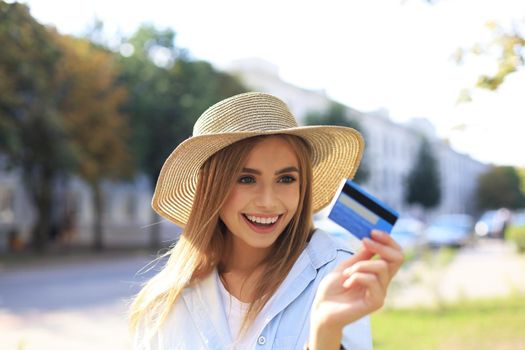 Cheerful young optimistic girl standing outdoors, holding credit card in hand.