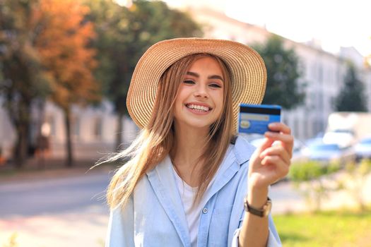 Cheerful young optimistic girl standing outdoors, holding credit card in hand.