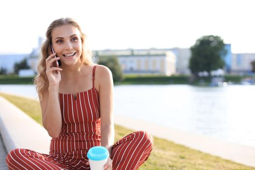 Young blonde woman sitting in riverbank outdoors talking by mobile phone.