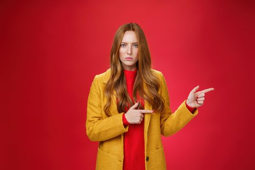 Angry and offended disappointed redhead female in yellow coat frowning looking strict and serious at camera with dislike and agression pointing right asking explain yourself over red background