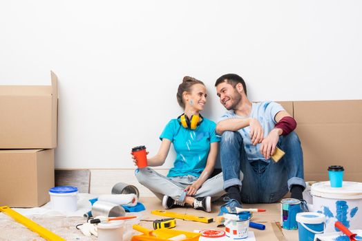 Happy boy and girl drinking coffee on floor. Home remodeling after moving. Cardboard boxes, construction tools and materials for building on floor. Couple having fun in time of house renovation.