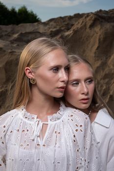 two young blond pretty twins posing at sand quarry in elegant white clothes