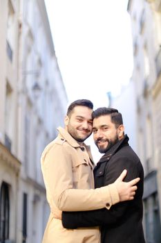 Two hugging handsome gays in city, buildings i background.
