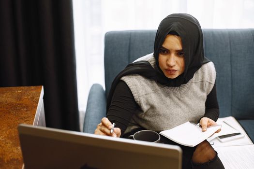 Arabic woman in headscarf using laptop at home, paying for utilities online.