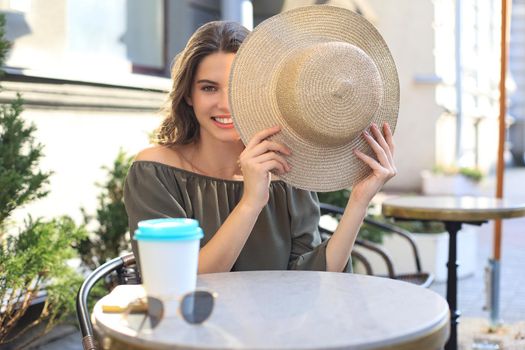 Portrait of a girl hiding her face behind a straw hat while sitting in summer street cafe.