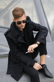 Young stylish man wearing black jacket sitting on Louvre Pyramid and smoking.