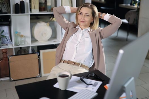 Woman with satisfied expression at desk. Arms up and folded behind her head in modern office.