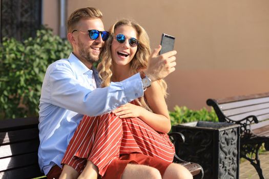 Romantic young couple in summer clothes smiling and taking selfie while sitting on bench in city street