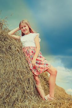 A beautiful blonde girl in a pink dress climbed on large bales of straw on a summer day