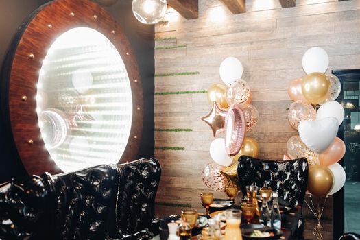 Black armchairs by the table and large illuminated mirror on the wall. Foiled balloons by the wall. Party concept