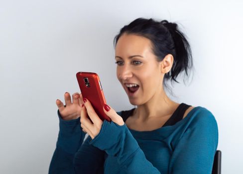 Young casual hipster woman holding and looking at cell phone reading unexpected sms news with a surprised and amazed facial expression, on white background